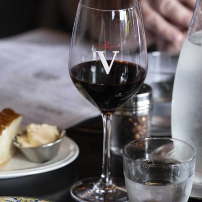 Glass of red wine with Valentina's V logo with focaccia and menu in background at Valentina's restaurant in Dublin, Ohio at Bridge Park.